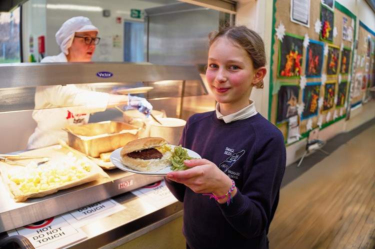 Girl poses in front of school dinner counter with wild venison burger. 